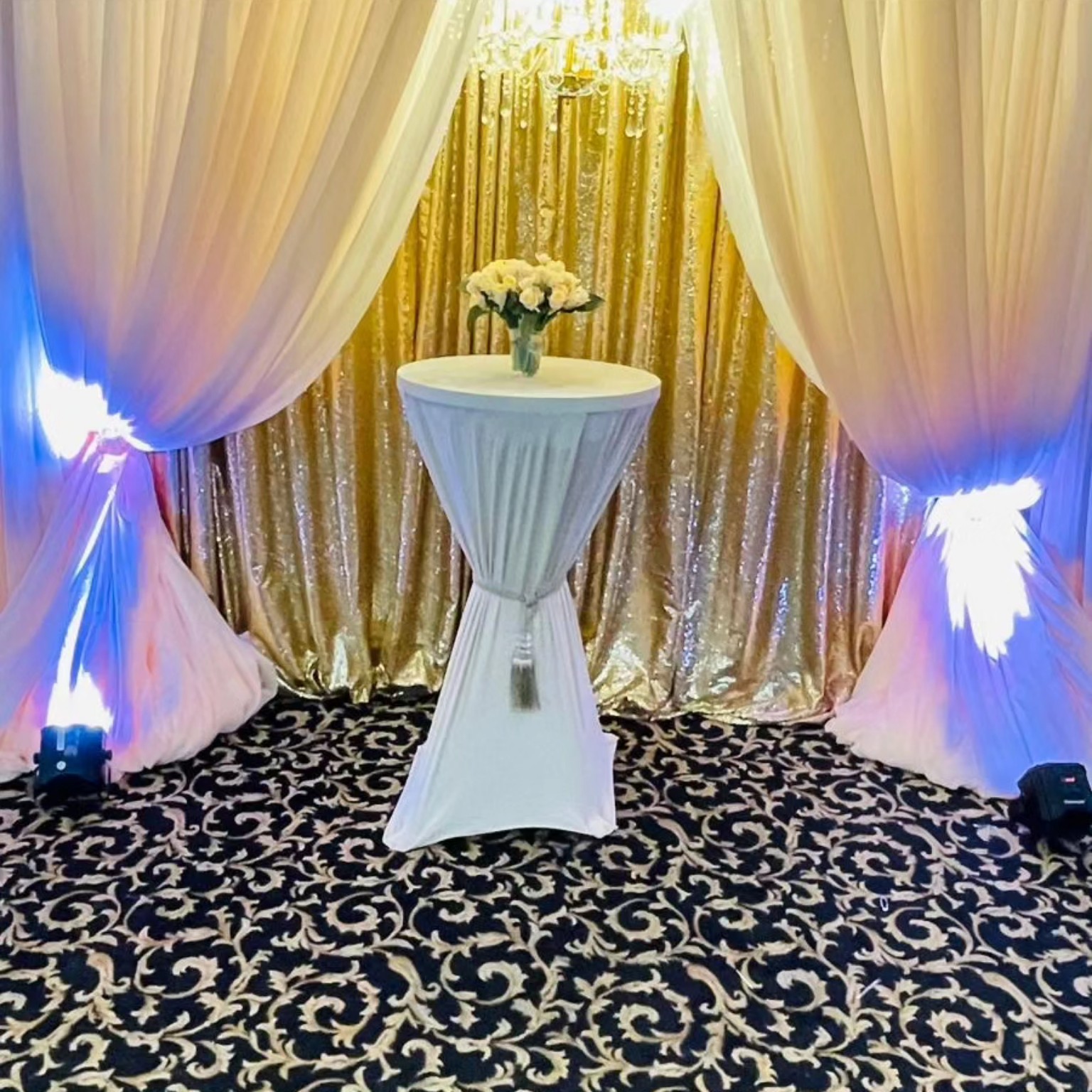 White draping and gold sequin backdrop framing a tall cocktail table with white flowers